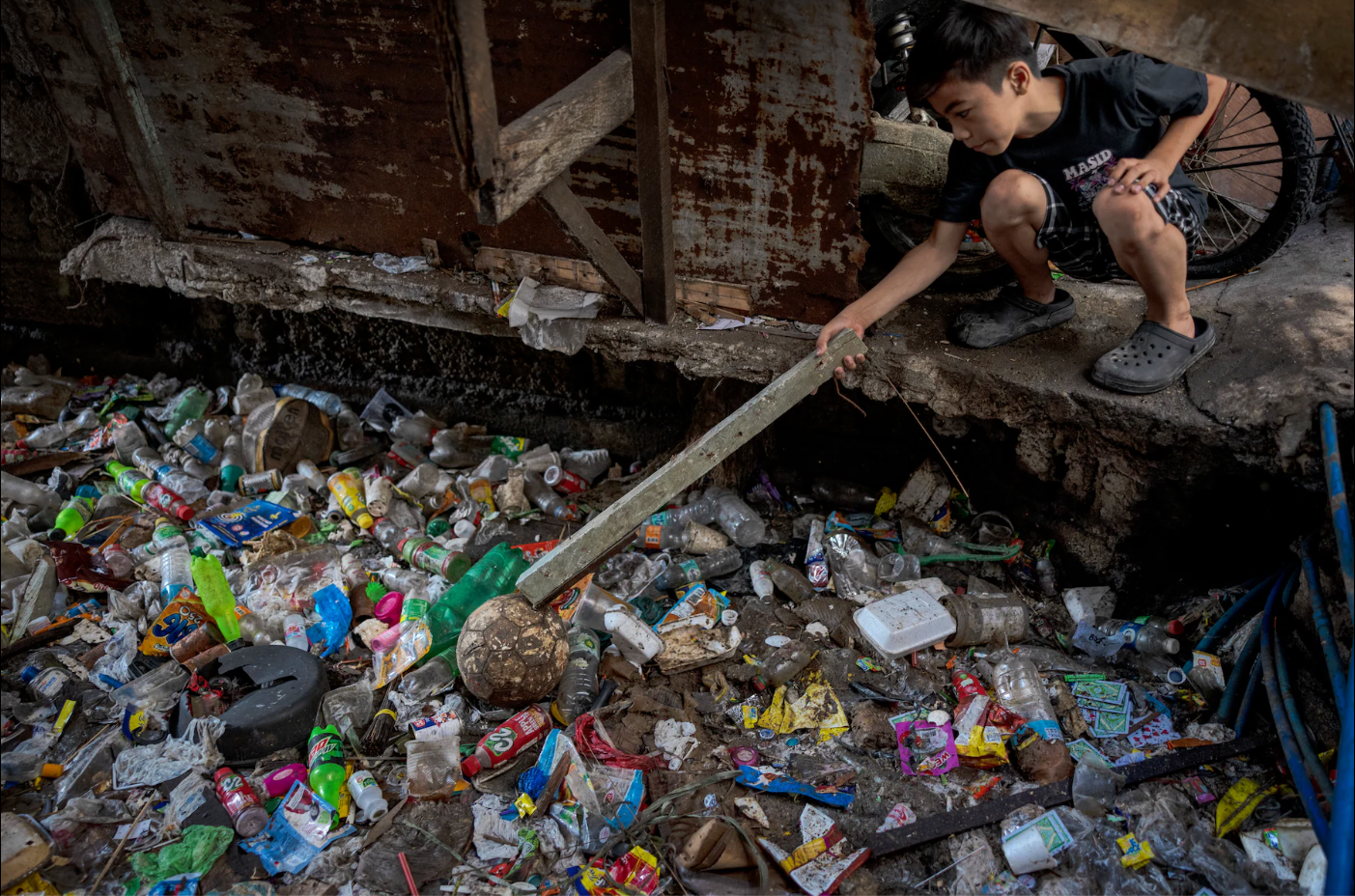 A young Filipino boy uses a wooden post to reach for his soccer ball that has rolled into a plastic landfill.