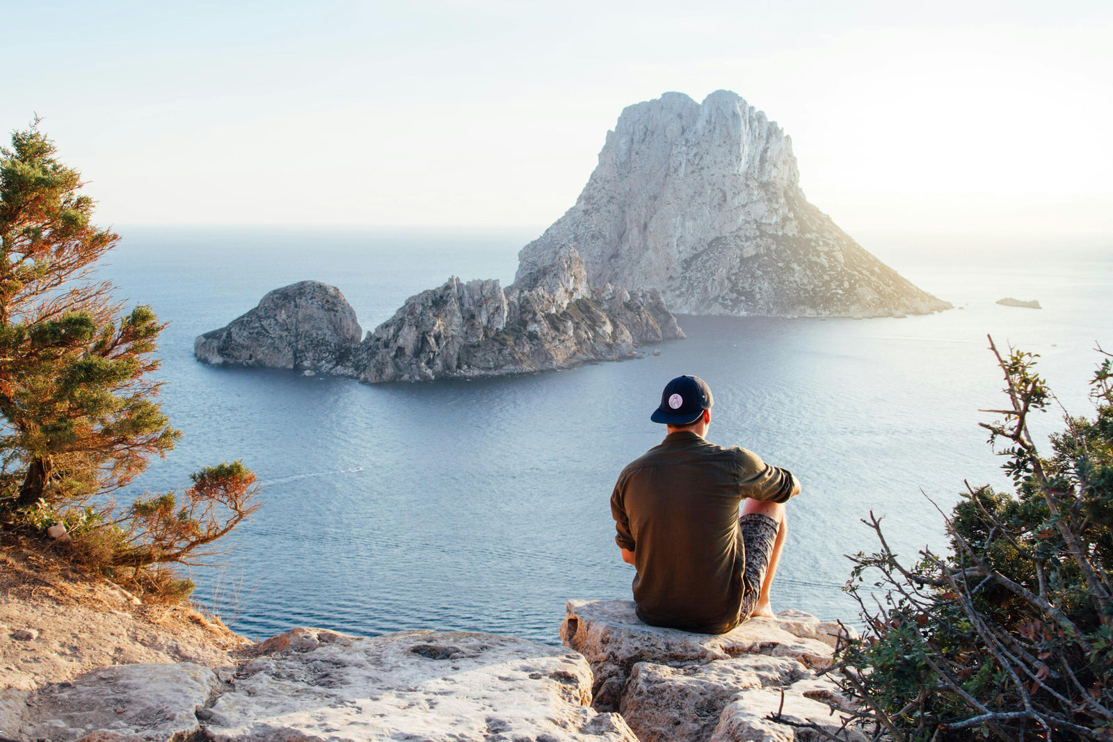 Man sitting on cliff, looking out at view of ocean and island. Sustainable travel with shampoo tablets. Eco-friendly adventure with family. 