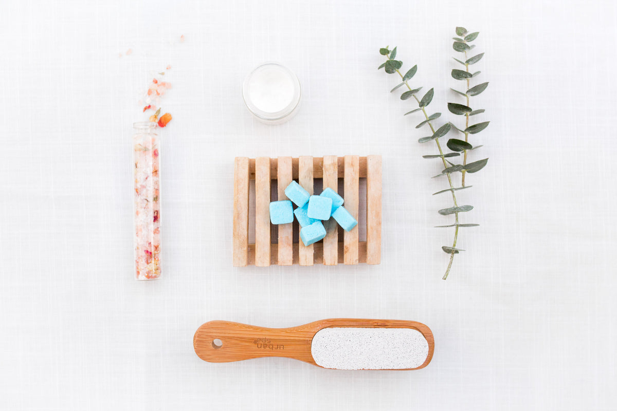 Image of EarthSuds travel shampoo, conditioner, body wash tablets sitting on wooden platform surrounded by shower brush, candle and plant